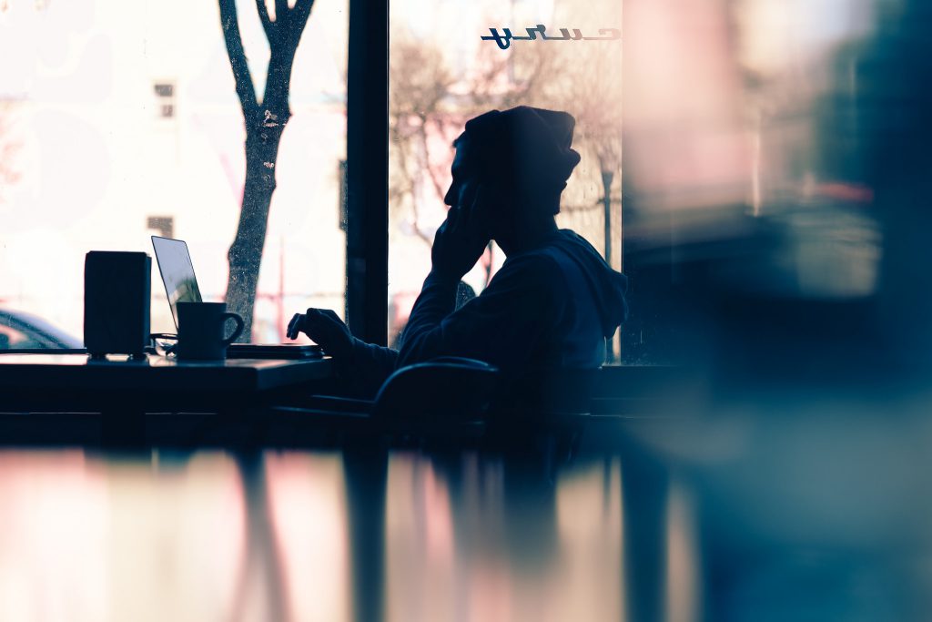 Backlit person wearing a toque sits at a table typing on a laptop, mug beside them